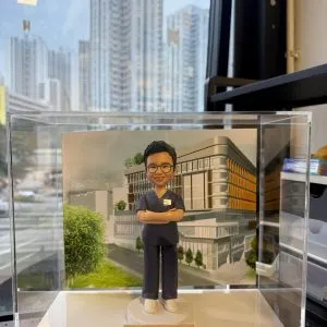 透明展示盒內的客製醫護公仔，穿深藍手術服雙手交叉站立，背板為現代醫學院建築，底座銘牌寫有「Dr. Veronica Lam School of Nursing, HKU」