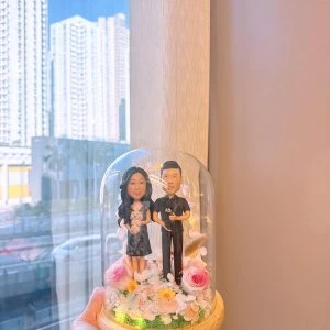 玻璃花盅內的一對客製情侶公仔，女方穿深色花裙抱白色小狗，男方穿黑色襯衫抱黑色小狗，周圍以粉玫瑰、白花與暖光燈點綴，木質底座襯草地