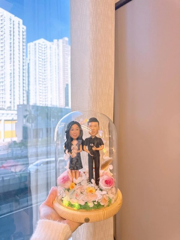 玻璃花盅內的一對客製情侶公仔，女方穿深色花裙抱白色小狗，男方穿黑色襯衫抱黑色小狗，周圍以粉玫瑰、白花與暖光燈點綴，木質底座襯草地