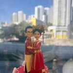 手持展示的客製中式婚禮情侶公仔，新郎穿紅金長衫，新娘著紅色秀禾服與華麗鳳冠，站在愛心形底座上