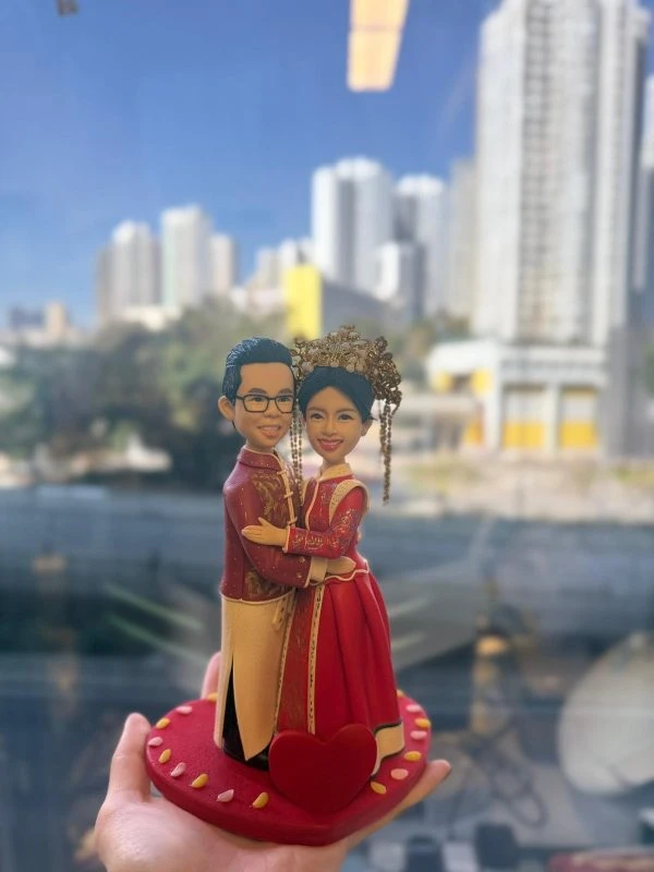 手持展示的客製中式婚禮情侶公仔，新郎穿紅金長衫，新娘著紅色秀禾服與華麗鳳冠，站在愛心形底座上