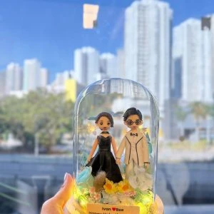 手持玻璃罩小夜燈式雙人公仔:木質圓形底座上放置透明玻璃罩,內有穿黑色禮服與白色西裝的公仔牽手站立,周圍點綴藍色與白色乾燥花、黃色小花與暖黃燈串;前方小名牌寫著「Ivan ♥ Bee Happy Valentine’s Day」。背景為窗外藍天與高樓城市景。