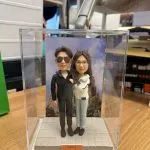 透明展示盒內的雙人客製公仔並肩站立，左側公仔穿深色外套與太陽眼鏡，右側公仔穿白色外套與淺色長褲；腳下為灰色方格地台，背板為歐式教堂風景照；前方橘色小名牌印有 2026.02.14 Valentine’s Day 字樣；擺放在木質桌面，背景為室內陳列與收納物品。