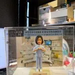 透明展示盒中的醫護主題客製公仔，穿著淺藍色護理服與白鞋，雙手在胸前比出愛心手勢；站在淺色小台座上，前方名牌寫有「APN Ms Leung Suk Yee Happy Retirement 2026」並配有紅色愛心；背板是新生兒加護病房設備與保溫箱的場景，展示盒上緣貼有「新生兒深切治療部及 新生兒特別護理部／Neonatal Intensive Care Unit & Special Care Baby Unit」字樣；背景為室內辦公環境。
