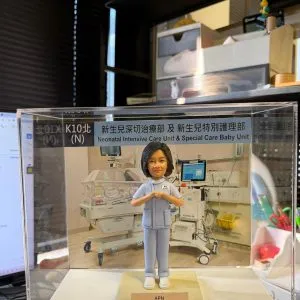 透明展示盒中的醫護主題客製公仔，穿著淺藍色護理服與白鞋，雙手在胸前比出愛心手勢；站在淺色小台座上，前方名牌寫有「APN Ms Leung Suk Yee Happy Retirement 2026」並配有紅色愛心；背板是新生兒加護病房設備與保溫箱的場景，展示盒上緣貼有「新生兒深切治療部及 新生兒特別護理部／Neonatal Intensive Care Unit & Special Care Baby Unit」字樣；背景為室內辦公環境。