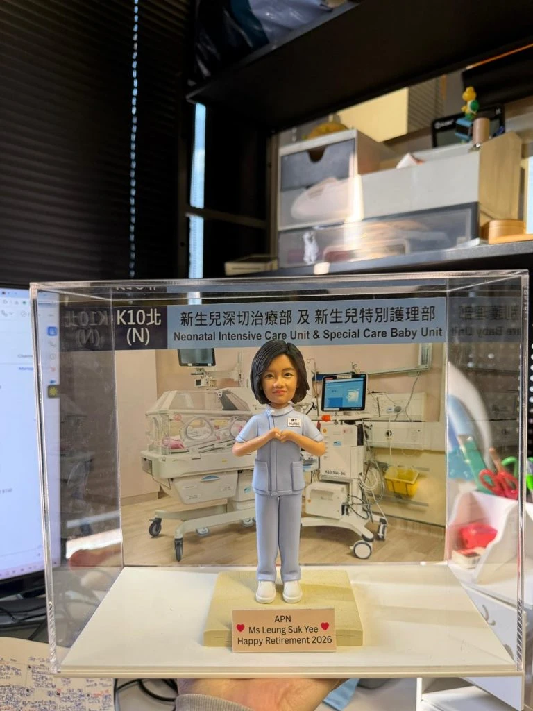 透明展示盒中的醫護主題客製公仔，穿著淺藍色護理服與白鞋，雙手在胸前比出愛心手勢；站在淺色小台座上，前方名牌寫有「APN Ms Leung Suk Yee Happy Retirement 2026」並配有紅色愛心；背板是新生兒加護病房設備與保溫箱的場景，展示盒上緣貼有「新生兒深切治療部及 新生兒特別護理部／Neonatal Intensive Care Unit & Special Care Baby Unit」字樣；背景為室內辦公環境。