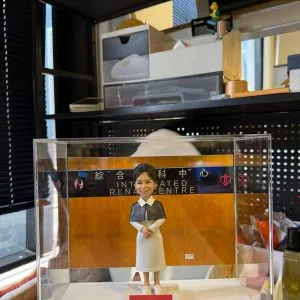 透明展示盒中的客製公仔,穿白色連身裙與深色披肩,雙手交握站在方形底座;背板為木質色並貼有「綜合腎科中心 INTEGRATED RENAL CENTRE」字樣與心形圖示,底座前有紅色小名牌寫著祝賀退休訊息;周圍可見辦公室置物架與雜物。