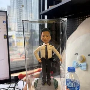 手持透明展示盒的近景照：盒內是一位身穿淺色制服襯衫、黑領帶與黑長褲的警務主題公仔，右手叉腰、左手持警帽；腳下為圓形灰色地磚基座，邊緣點綴綠地與小花紋；背景為辦公室桌面與窗外高樓，右側可見一瓶礦泉水。